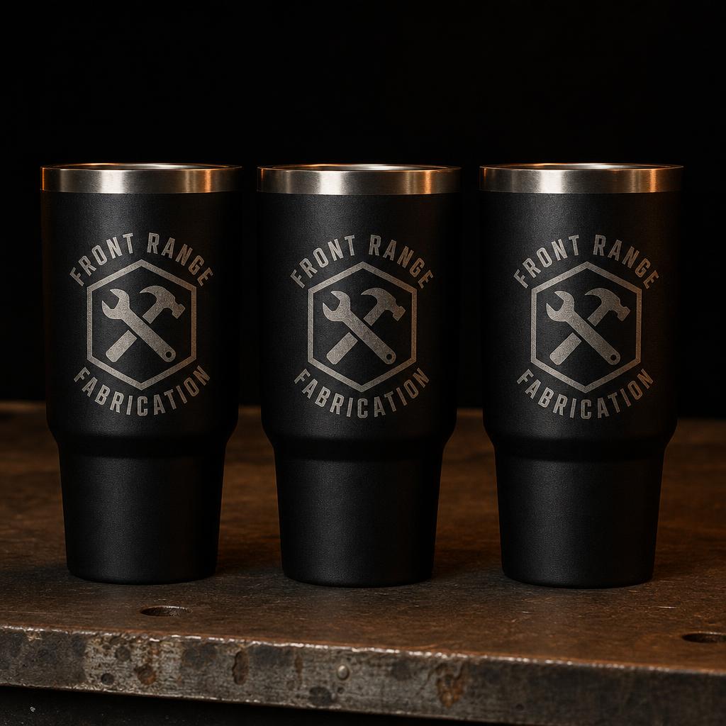 Row of black anodized steel tumblers laser-engraved with an industrial logo on a steel bench